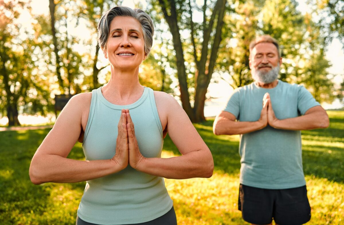Smiling senior couple closing eyes and doing yoga exercises.