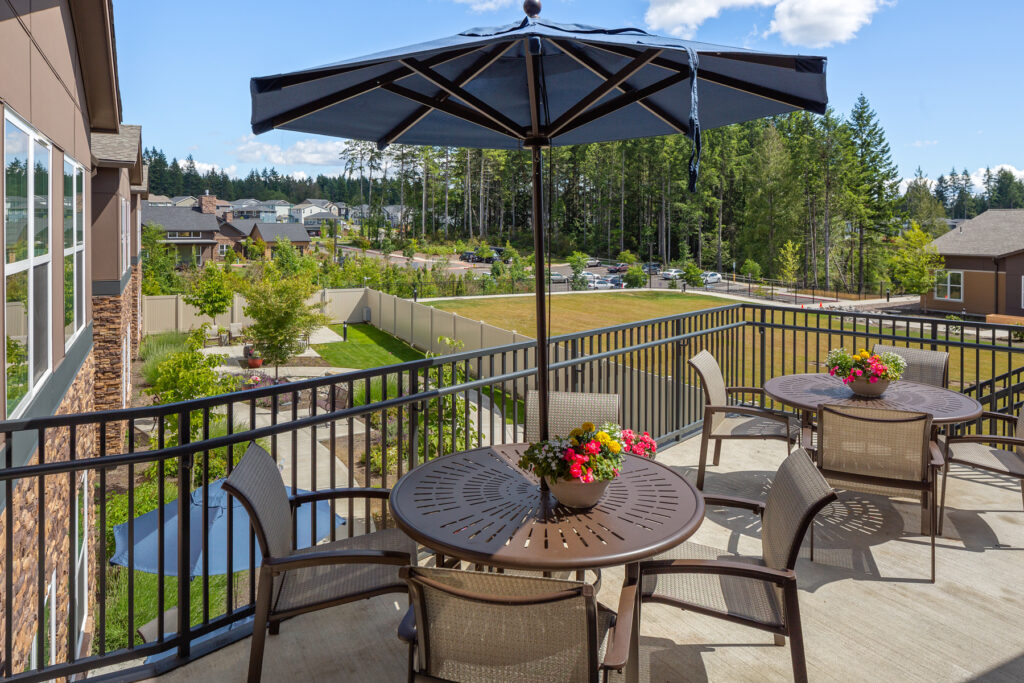 A balcony with four chairs and tables, shaded by an umbrella, overlooking a lush green landscape with trees and homes in the distance on a sunny day.