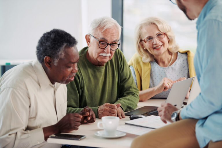 Older adults gathered around a table, discussing information on a tablet during a small group learning conversation.