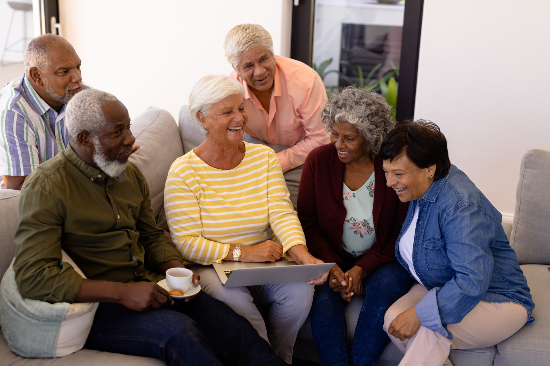 Multiracial senior woman holding laptop laughing while sitting with friends on sofa in retirement community